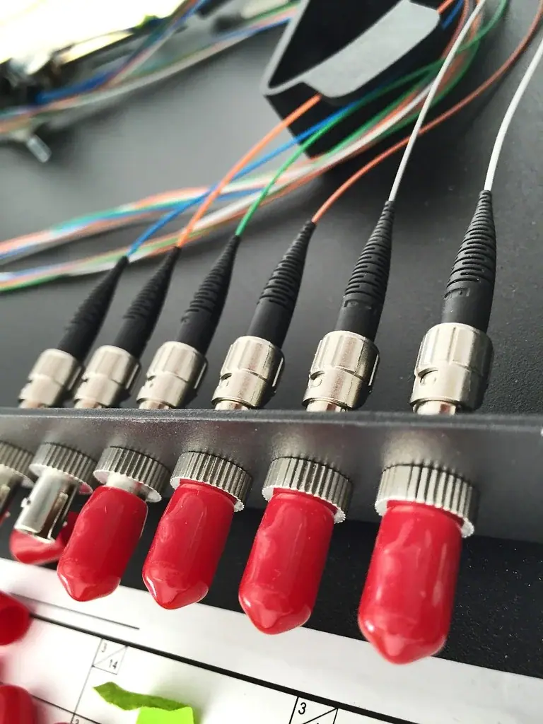 Close-up of metal-bodied fibre optic connectors with coloured pigtail fibres in a distribution frame, with red dust caps protecting the unmated bulkhead ports on the lower row